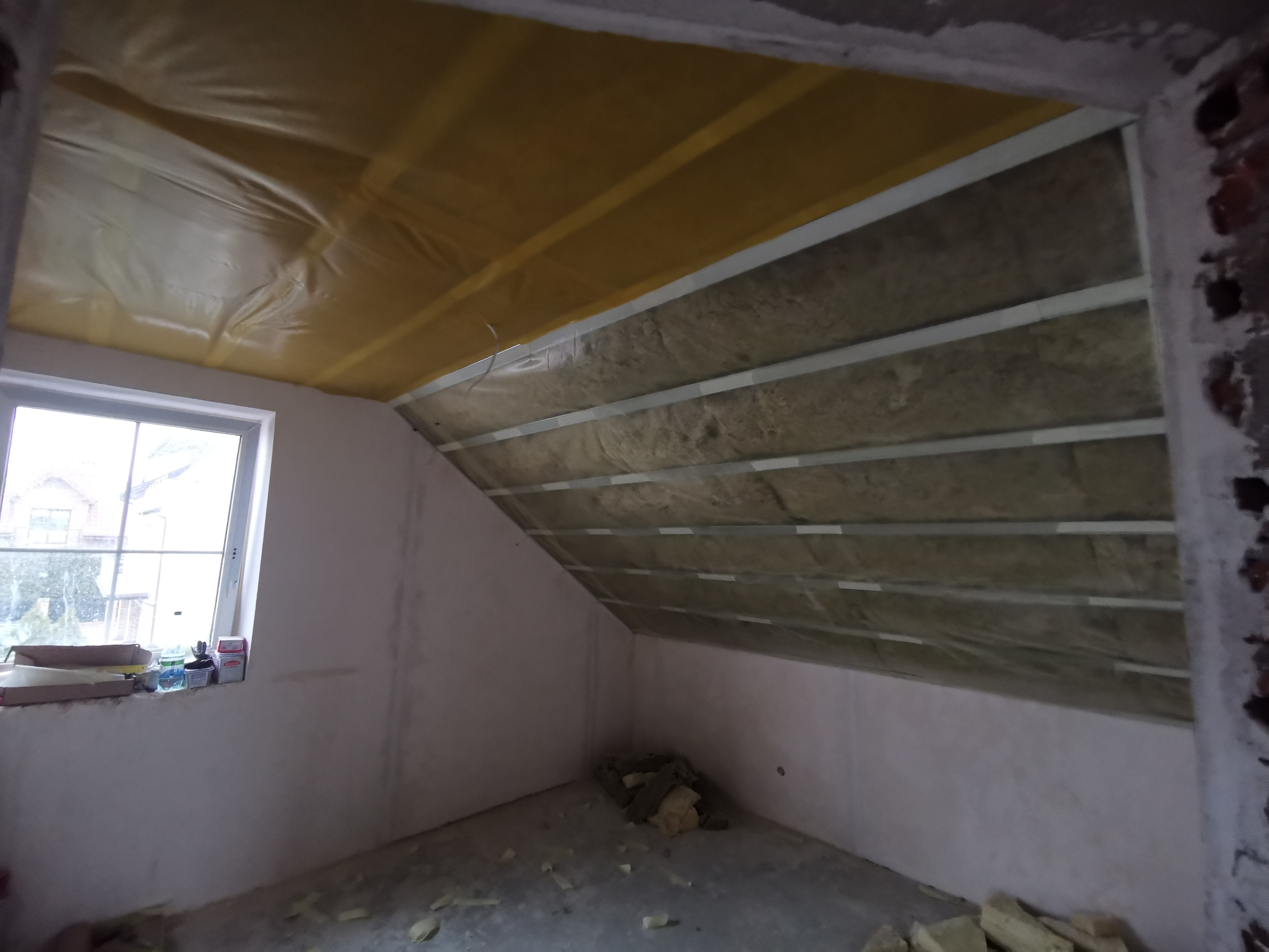 Poddasze w trakcie ocieplania wełną mineralną, widoczne profile metalowe, folia paroizolacyjna i fragment muru z cegły.