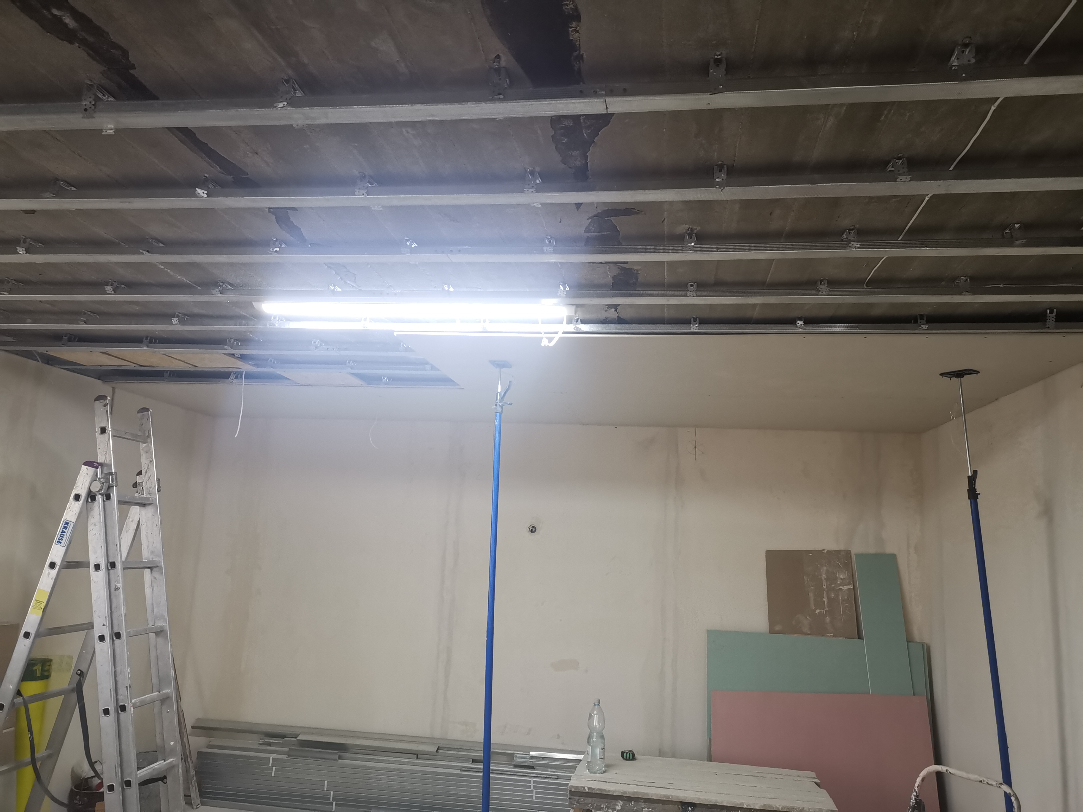 Montaż stelażu sufitu podwieszanego: widoczne profile CD zamocowane do sufitu, drabina Krause, podpory teleskopowe i płyty kartonowo-gipsowe w tle.