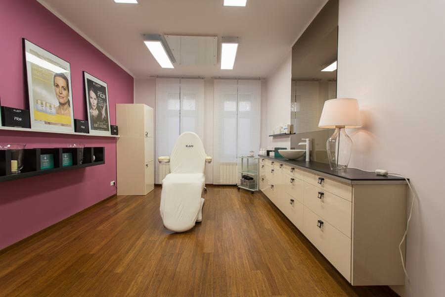 Jasne wnętrze gabinetu kosmetycznego z fotelem zabiegowym przykrytym białym pokrowcem z logo Klapp, umywalką nablatową i plakatami kosmetyków na różowej ścianie.