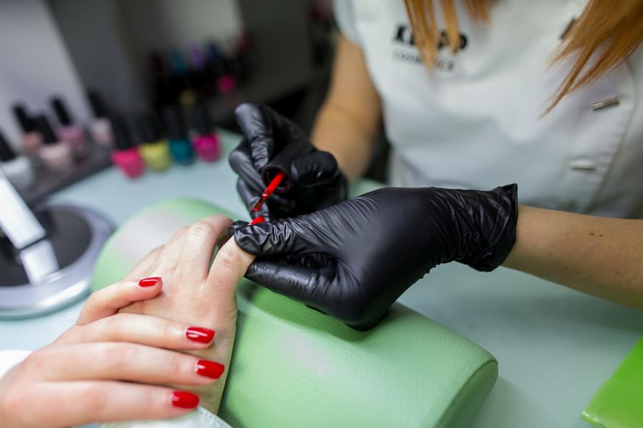 Nakładanie czerwonego lakieru na paznokcie dłoni, leżącej na zielonym podłożu, przez stylistkę w czarnych rękawiczkach, z widocznymi buteleczkami lakierów w tle.