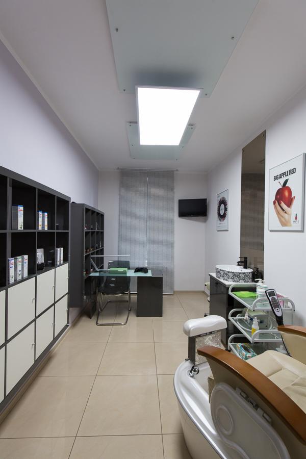 Wnętrze gabinetu z fotelem do pedicure, stolikiem do manicure, regałami z produktami i plakatem reklamowym OPI Big Apple Red.