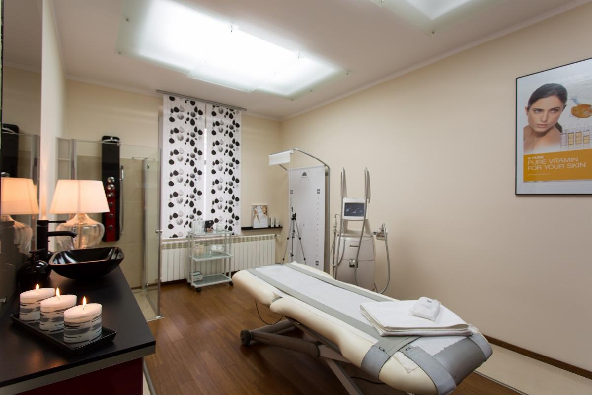 Jasne wnętrze gabinetu z łóżkiem zabiegowym, aparaturą kosmetyczną, czarną umywalką i płonącymi świecami, akcentowane zasłonami w czarno-białe grochy i plakatem reklamowym.