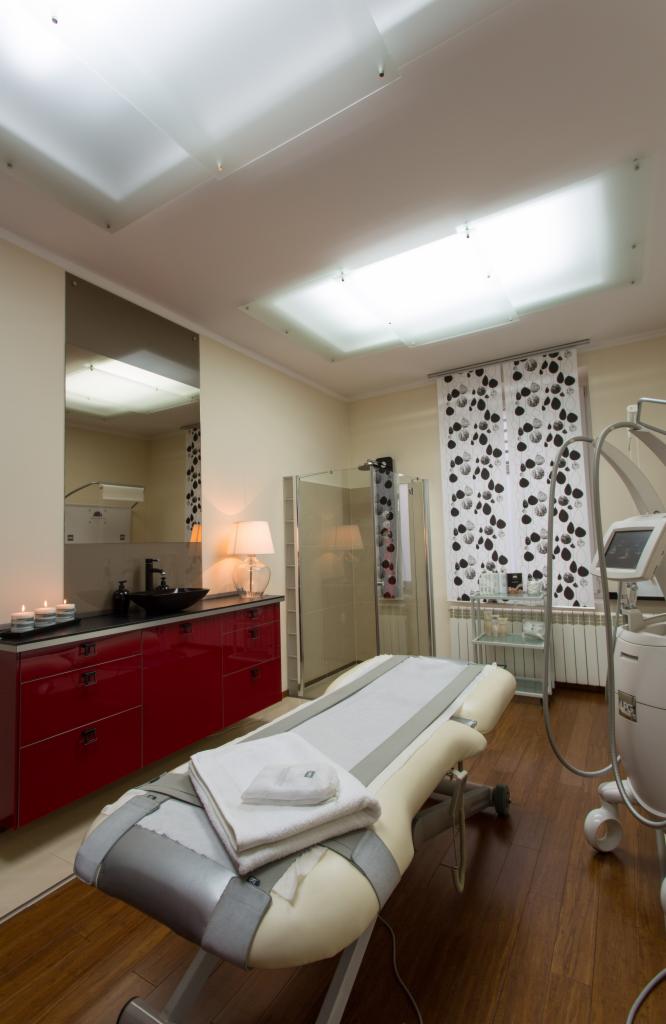 Jasne wnętrze gabinetu z łóżkiem do zabiegów z ręcznikami, czerwona szafka z umywalką, kabina prysznicowa i urządzenie do modelowania sylwetki.