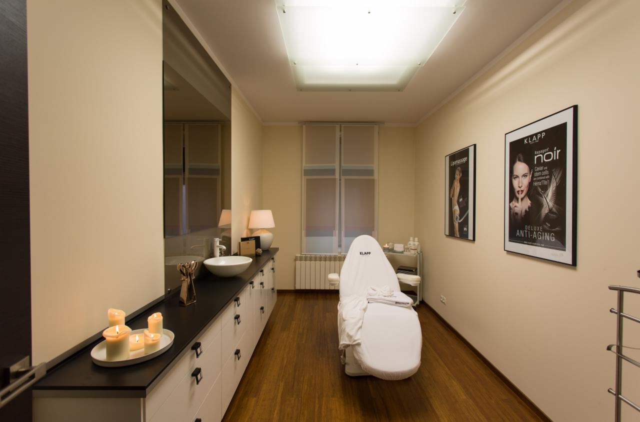 Jasne wnętrze gabinetu z fotelem kosmetycznym przykrytym białym pokrowcem z logo KLAPP, umywalką na blacie z zapalonymi świecami, plakatami na ścianie i drewnianą podłogą.