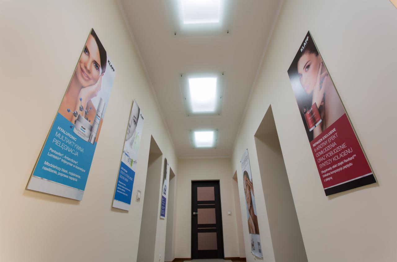 Korytarz gabinetu z plakatami reklamowymi kosmetyków marki Klapp na ścianach, podłużne oświetlenie sufitowe.