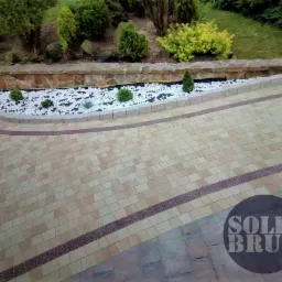 Beżowa kostka brukowa ułożona w łuk, obrzeże z białych kamyków i niskie rośliny, logo firmy w prawym dolnym rogu.