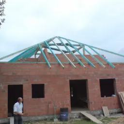 Wznoszony dom murowany z częściowo wykonaną, pomalowaną na niebiesko więźbą dachową; widoczny pracownik, narzędzia i beczka z napisem SOPUR.