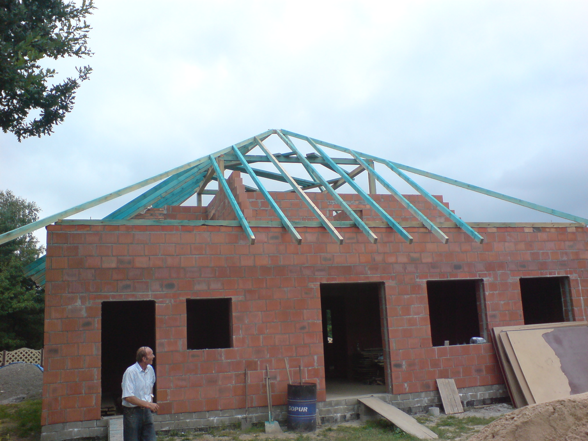 Wznoszony dom murowany z częściowo wykonaną, pomalowaną na niebiesko więźbą dachową; widoczny pracownik, narzędzia i beczka z napisem SOPUR.