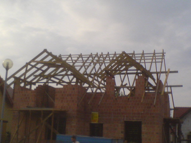 Budowa domu z czerwonej cegły, widoczna drewniana konstrukcja dachu w trakcie montażu, słabe oświetlenie.