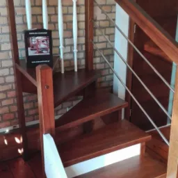 Drewniane schody o ciemnym odcieniu z białymi podstopnicami i balustradą z elementami ze stali nierdzewnej, w tle ceglana ściana i katalog schodów na półce.