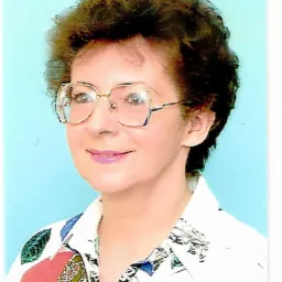 Portret kobiety w okularach o złotych oprawkach, z krótkimi kręconymi włosami, ubrana w białą koszulę z kolorowym wzorem na jasnoniebieskim tle.