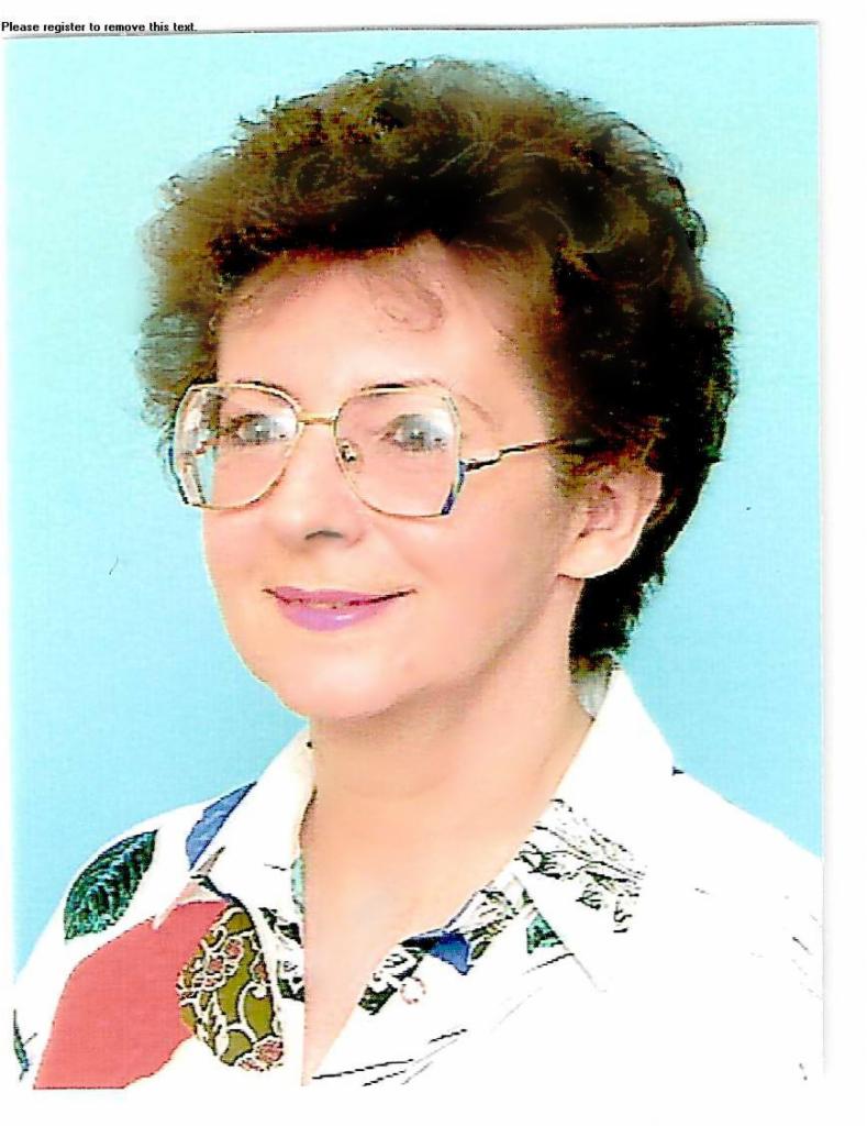 Portret kobiety w okularach o złotych oprawkach, z krótkimi kręconymi włosami, ubrana w białą koszulę z kolorowym wzorem na jasnoniebieskim tle.