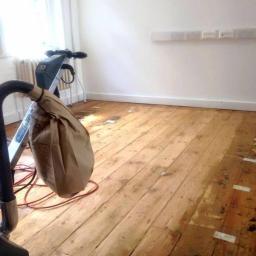 Abc Rusztowania Dawid Saganowski - Szlifowanie drewnianej podłogi w pomieszczeniu z białymi ścianami, widoczna maszyna do szlifowania i częściowo odnowiona powierzchnia.