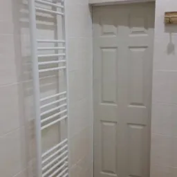 Wnętrze łazienki z białym grzejnikiem drabinkowym, beżowymi płytkami ściennymi i podłogą imitującą drewno, widoczna biała, sześciopanelowa  drzwi.