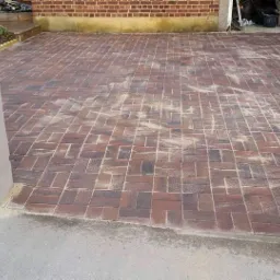 Nowo ułożona, brązowo-czerwona kostka brukowa na podjeździe, z zagęszczarką w tle i śladami piasku na powierzchni.