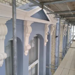 Elewacja budynku w trakcie malowania, widoczne rusztowania i detale architektoniczne, świeża warstwa farby w odcieniach szarości i beżu.