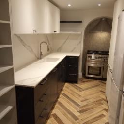 Abc Rusztowania Dawid Saganowski - Nowoczesna kuchnia z białymi szafkami górnymi, ciemnymi szafkami dolnymi z podłużnymi uchwytami, marmurowym blatem i backsplashiem, oraz podłogą ułożoną w jodełkę. W tle widoczny piekarnik w niszy.