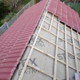 Abc Rusztowania Dawid Saganowski - Częściowo pokryty czerwony dach z widoczną konstrukcją drewnianą i membraną dachową z nadrukiem, widok z góry na ogród z budynkami gospodarczymi.