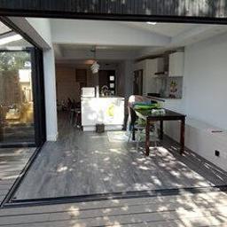 Abc Rusztowania Dawid Saganowski - Otwarta przestrzeń: szare panele podłogowe przechodzące z tarasu do wnętrza domu z widoczną kuchnią i jadalnią przez rozsuwane, czarne, panoramiczne drzwi.