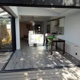 Otwarta przestrzeń: szare panele podłogowe przechodzące z tarasu do wnętrza domu z widoczną kuchnią i jadalnią przez rozsuwane, czarne, panoramiczne drzwi.