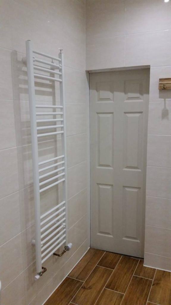 Wnętrze łazienki z białym grzejnikiem drabinkowym, beżowymi płytkami ściennymi i podłogą imitującą drewno, widoczna biała, sześciopanelowa  drzwi.