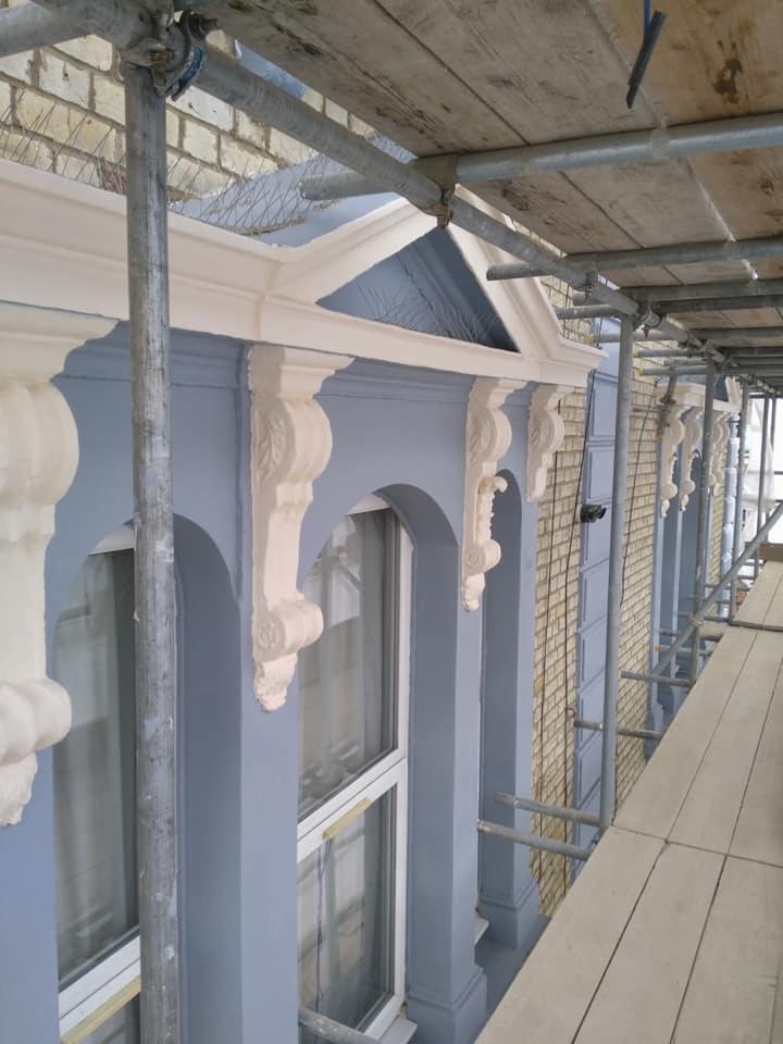 Elewacja budynku w trakcie malowania, widoczne rusztowania i detale architektoniczne, świeża warstwa farby w odcieniach szarości i beżu.