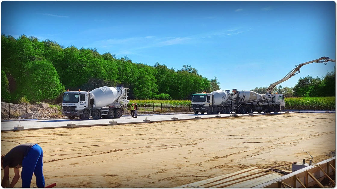 Budowa hali w Bydgoszczy: betonowanie fundamentów pod konstrukcję. Dwa betonomieszarki i pracownicy na placu budowy. Widok na teren inwestycji.