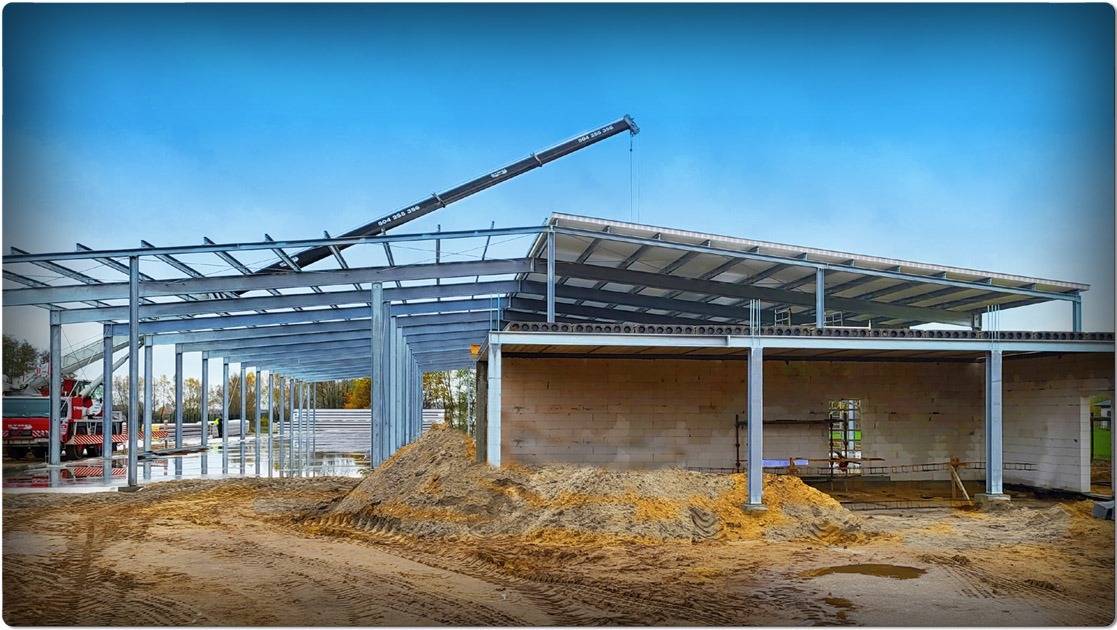 Wznoszenie hali: stalowa konstrukcja szkieletowa z dźwigiem na tle błękitnego nieba. Widoczne bloczki fundamentowe i ziemia. Etap budowy.