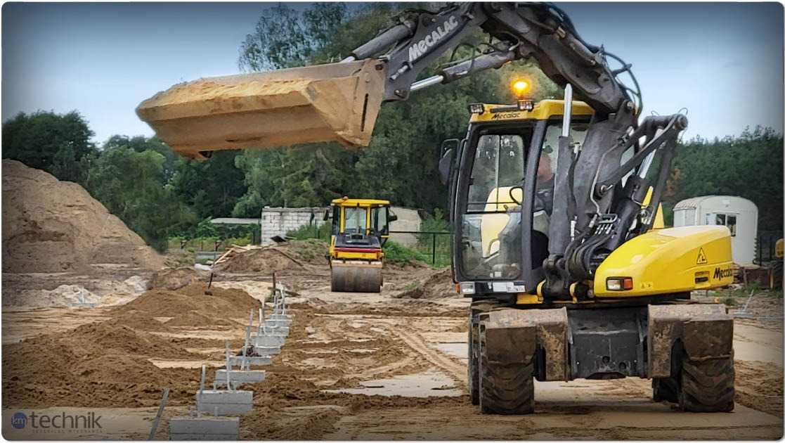 Żółta koparka Mecalac z łyżką uniesioną nad fundamentami hali, w tle walec drogowy i hałda piasku. Etap przygotowania terenu pod budowę.