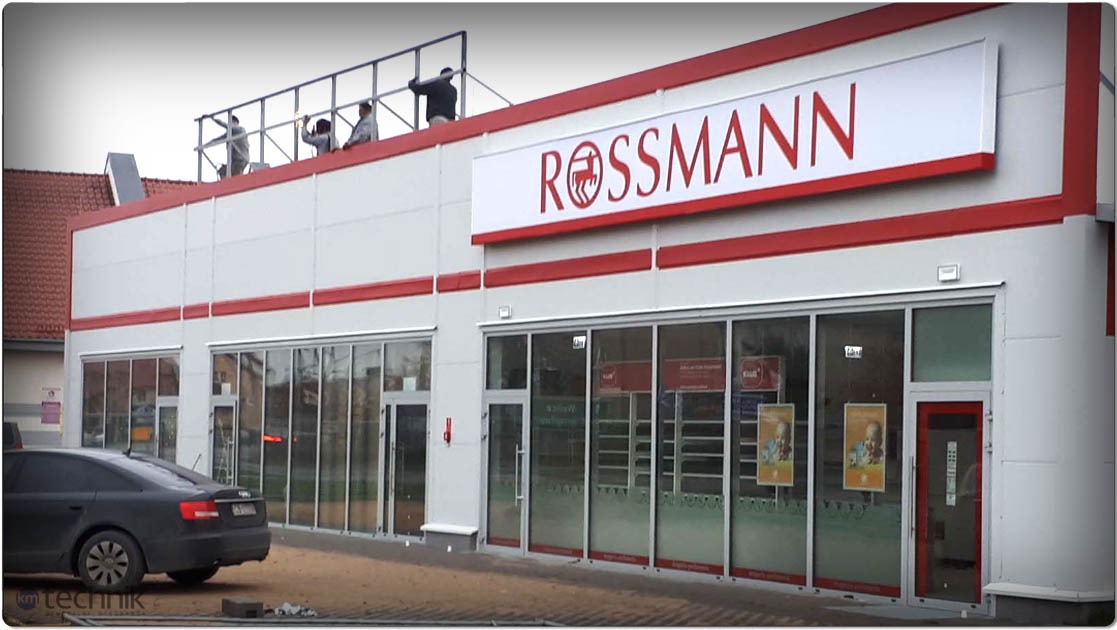 Nowoczesna hala handlowa Rossmann z przeszkloną fasadą i czerwonymi akcentami. Na dachu pracownicy montują konstrukcję. Zaparkowany samochód osobowy.