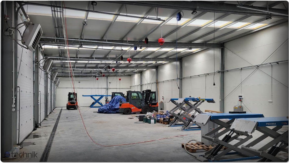Wnętrze hali z konstrukcją stalową, oświetleniem i bramami segmentowymi. Widoczne podnośniki samochodowe i wózki widłowe. Hala w trakcie wyposażania.