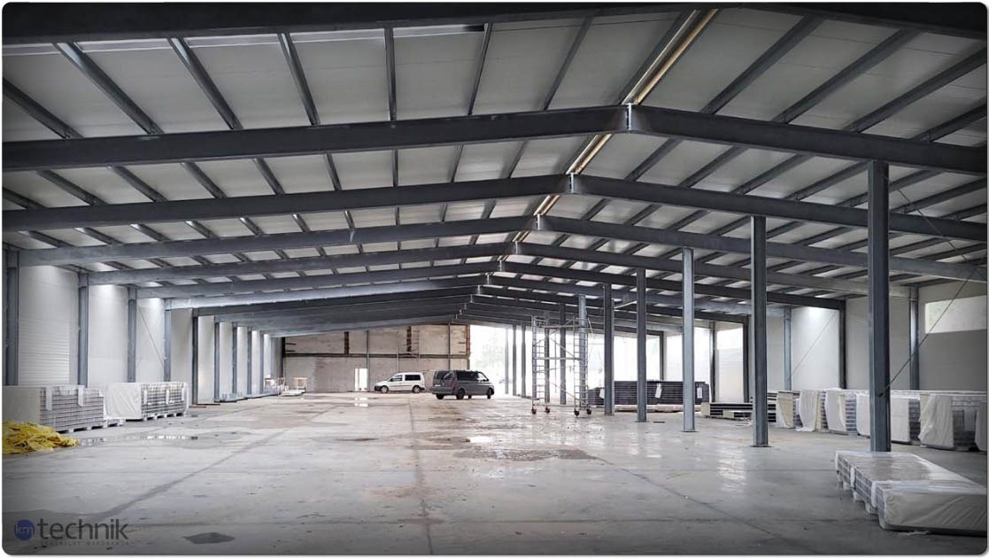 Wnętrze hali w trakcie budowy. Widoczne stalowe elementy konstrukcyjne, częściowo zamontowane płyty stropowe, materiały budowlane i dwa samochody w tle. Surowy beton na podłodze.