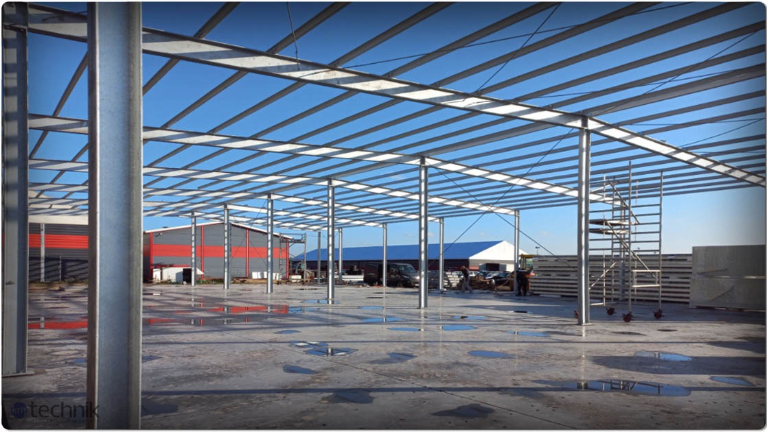 Wnętrze hali w budowie: stalowa konstrukcja szkieletowa, kolumny, kratownica dachu, kałuże na posadzce, w tle budynki i drabina. Błękitne niebo widoczne przez konstrukcję.