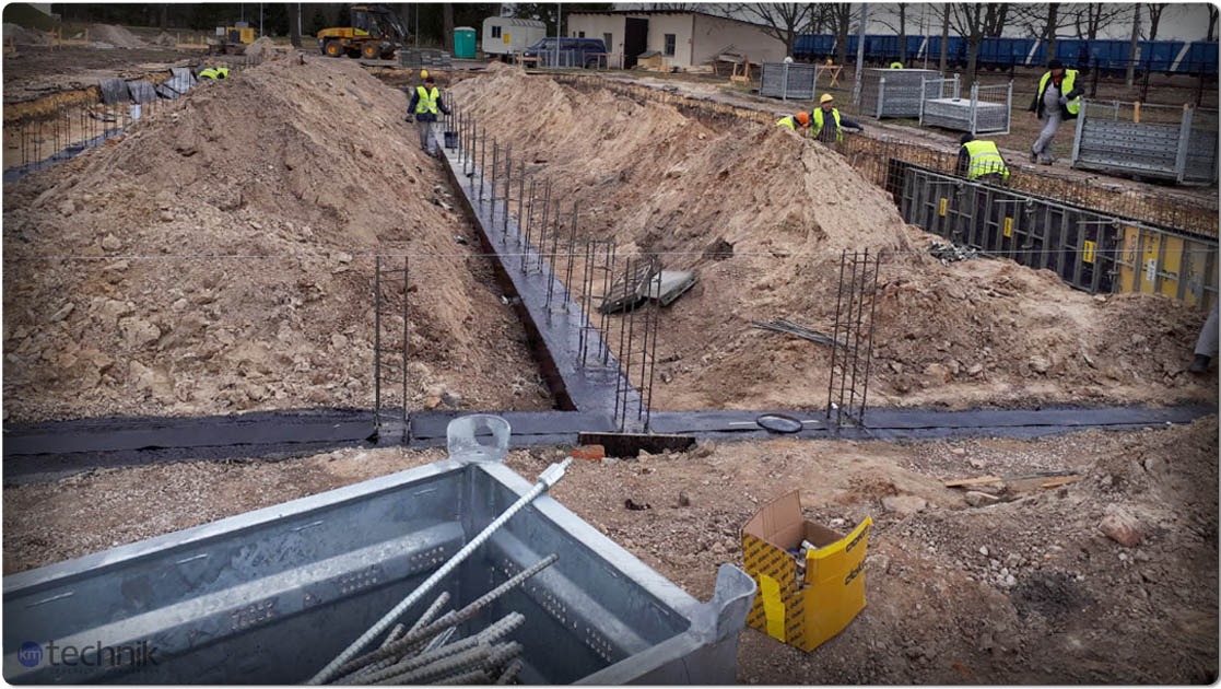Fundamenty hali w trakcie budowy: wykop zbrojony stalą, widoczni pracownicy w odblaskowych kamizelkach, ciężki sprzęt w tle. Ujęcie z bliska na zbrojenie.