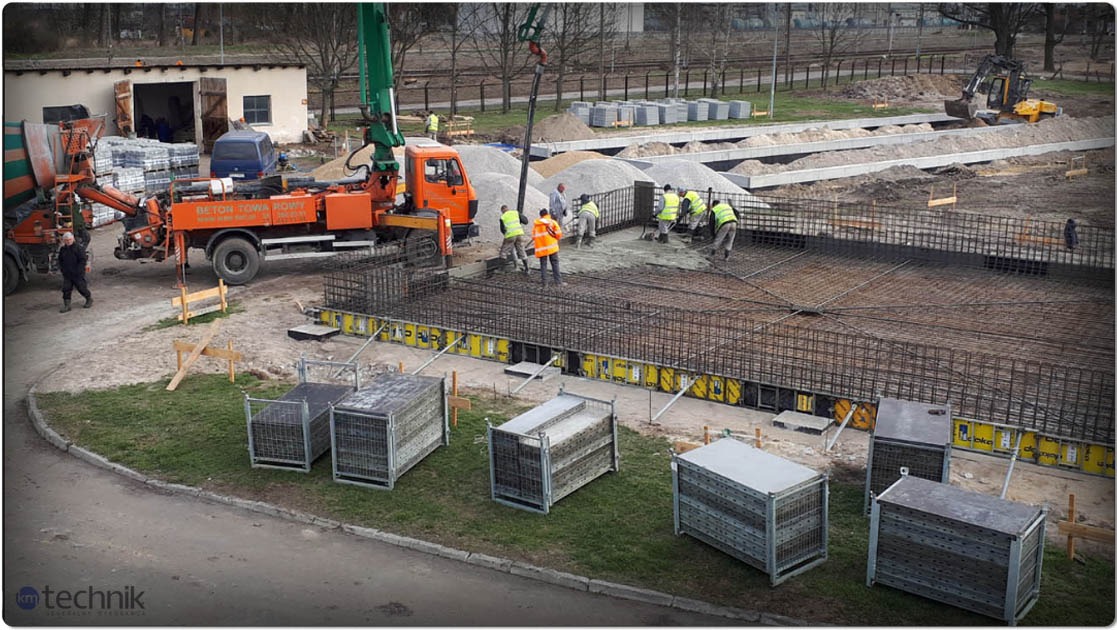 Wylewanie betonu na zbrojenie fundamentów hali. Widoczny betonowóz z pompą, pracownicy w odblaskowych kamizelkach i szalunki. W tle budynek i teren budowy.