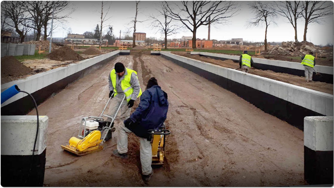 Prace ziemne przy budowie hali: dwóch robotników zagęszcza podłoże ubijakami wibracyjnymi. W tle widoczne fundamenty i materiały budowlane.