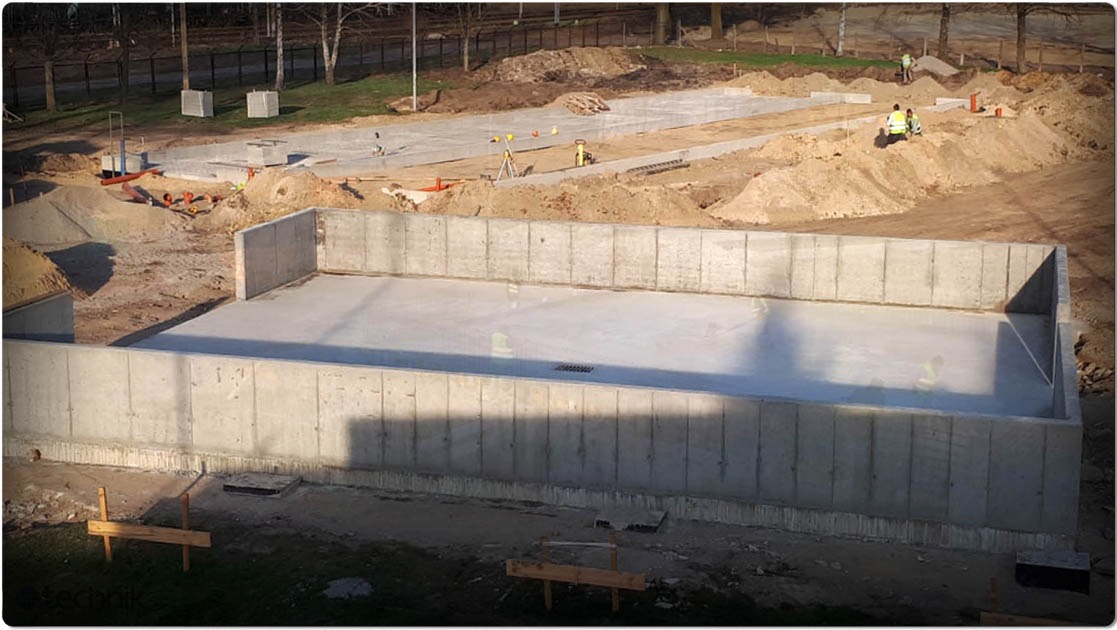 Wylana płyta fundamentowa pod halę z betonowymi ścianami oporowymi. W tle widoczne przygotowane podłoże pod kolejne etapy budowy. Prace ziemne w toku.