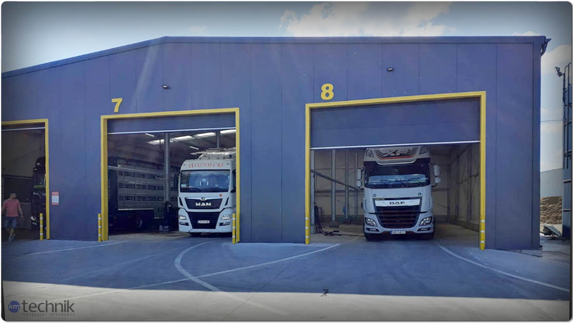 Nowoczesna hala magazynowa z numerami 7 i 8 nad bramami, z wjeżdżającymi ciężarówkami MAN i DAF. Żółte obramowania bram, szara elewacja. Widoczny fragment nieba.