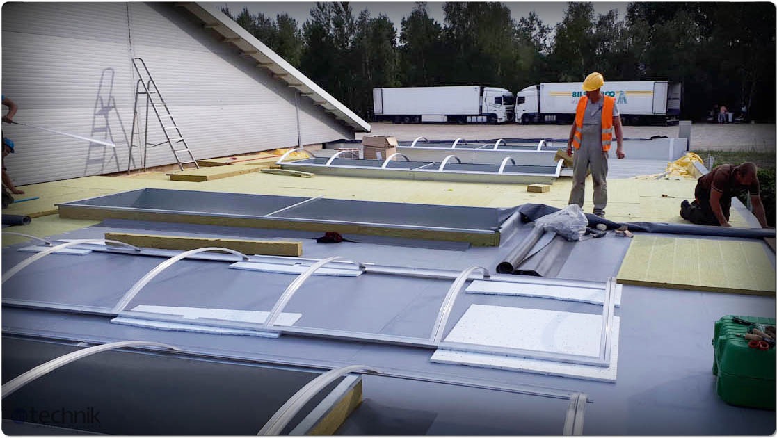 Montaż świetlików dachowych na hali. Pracownicy w kaskach i kamizelkach odblaskowych układają izolację i elementy konstrukcyjne. W tle zaparkowane ciężarówki.