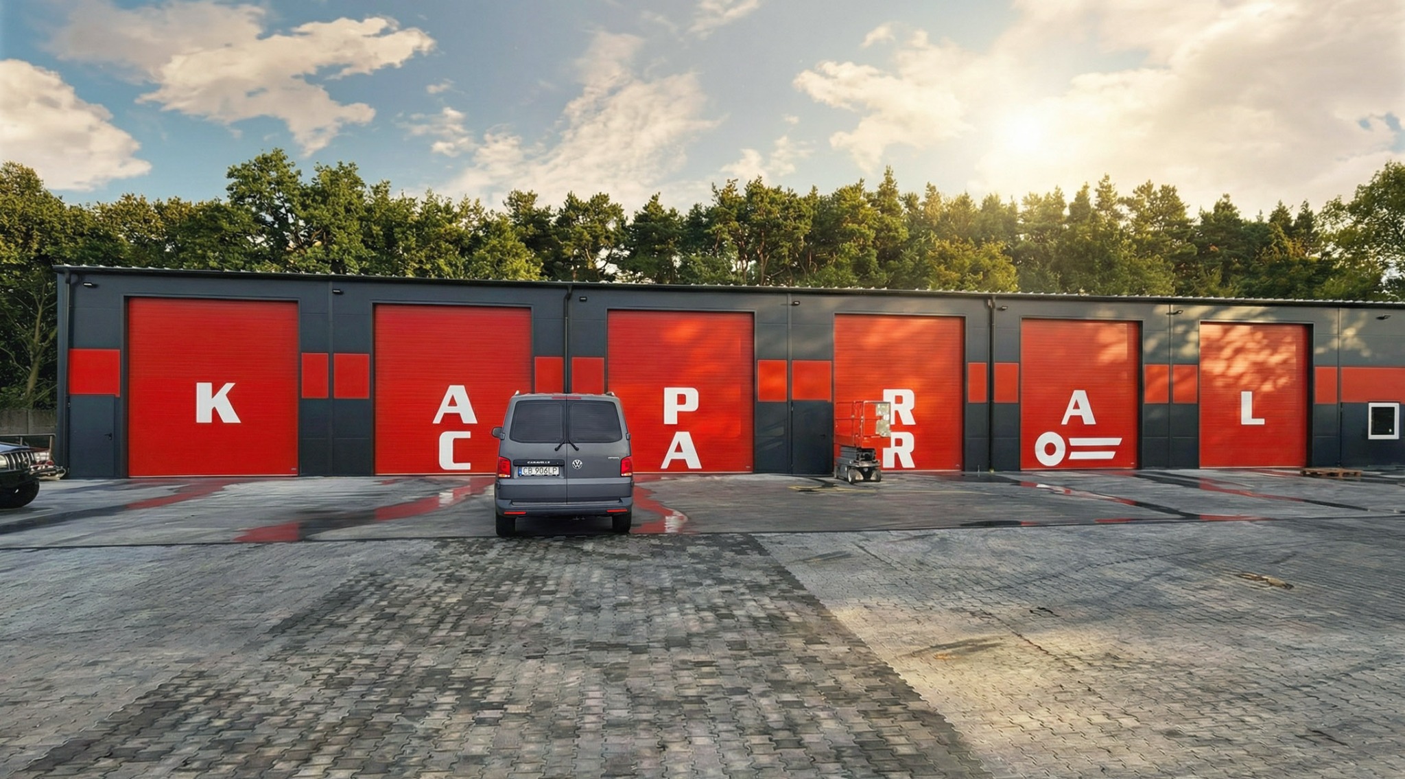 Nowoczesna hala z czerwonymi bramami segmentowymi, na których widnieje napis 'KACPARAL'. Szara kostka brukowa i zaparkowany bus. Słoneczny dzień.
