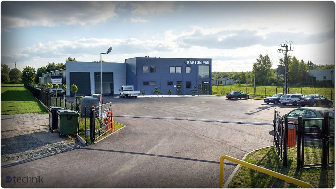 Nowoczesna hala produkcyjna Karton Pak z biurem, parkingiem i zielenią. Widok z zewnątrz, brama wjazdowa, szara elewacja, błękitne akcenty, słoneczny dzień.