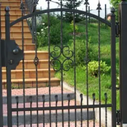 Ciemna, metalowa brama z dekoracyjnymi elementami na tle schodów i zieleni ogrodu, skrzynka na listy po lewej stronie.