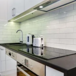 Biała kuchnia z czarnym blatem, podświetleniem LED podszafkowym, białymi płytkami na ścianie, zlewem, czajnikiem i tosterem