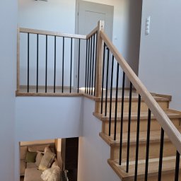FRED P.P.U - Jasne drewniane schody z czarnymi, metalowymi balustradami w nowoczesnym wnętrzu. Widok z góry na salon z sofą. Minimalistyczny design.