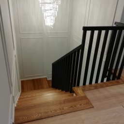 FRED P.P.U - Drewniane schody z czarną, pionową balustradą w nowoczesnym wnętrzu z dekoracyjną ścianą i kryształowym żyrandolem, widok z góry.