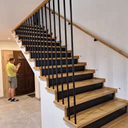 FRED P.P.U - Nowoczesne schody drewniane z czarnymi, metalowymi balustradami i jasną poręczą. Mężczyzna robi zdjęcie schodów telefonem. Minimalistyczny design, jasne wnętrze.