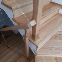 Jasne, drewniane stopnie z balustradą ze szkła i drewnianą poręczą, wspartą na złotych, metalowych elementach.