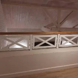 Biała balustrada z drewnianą poręczą na antresoli, wzór krzyżowy, białe ściany z cegły i drewniany sufit w tle.