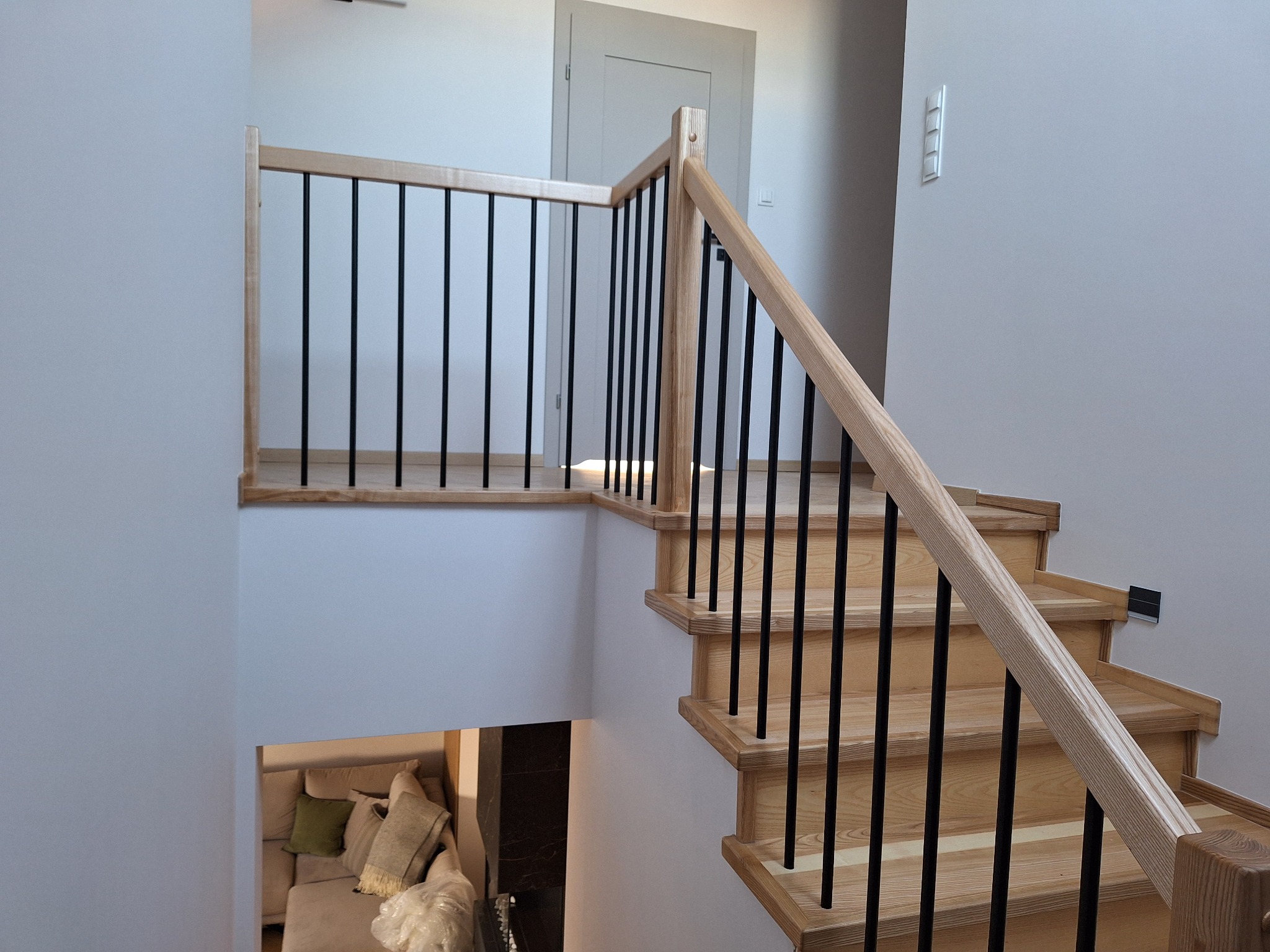 Jasne drewniane schody z czarnymi, metalowymi balustradami w nowoczesnym wnętrzu. Widok z góry na salon z sofą. Minimalistyczny design.