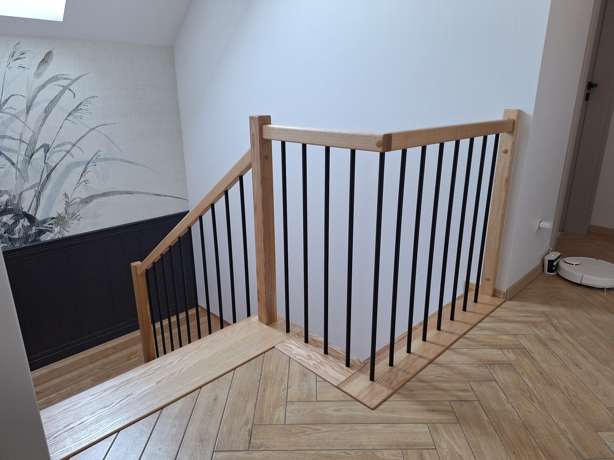 Drewniana balustrada schodowa z czarnymi, pionowymi szczeblami, widoczna z góry, na tle parkietu ułożonego w jodełkę i ściany z motywem roślinnym.